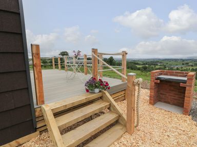 A wooden deck with rope railing, metal chairs and table, flowers, stairs leading down, and a brick barbecue with countryside view at Laird Lodge 1 in Lochmaben