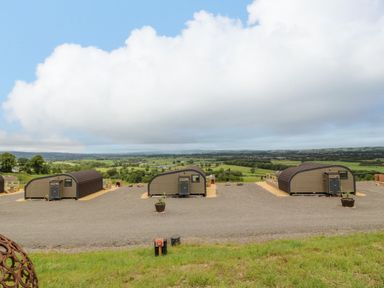 Three small arched cabins on a gravel area with green fields and hills in the background at Laird Lodge 1 in Lochmaben