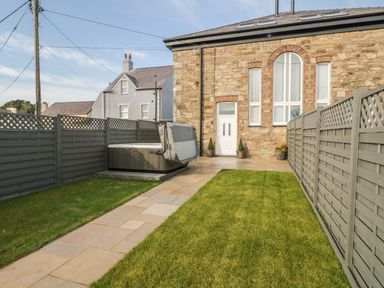 A fenced backyard with grassy areas and a paved path leading to a door on a stone building with large windows at Sardis Chapel 1 Malltraeth
