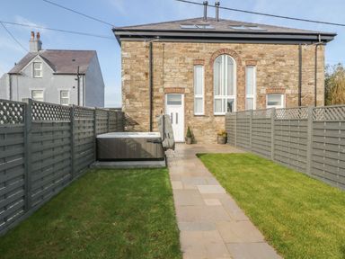 A backyard with a stone pathway leading to a stone house with tall windows and a door fenced by gray wooden panels and a hot tub on the left side at Sardis Chapel 1 Malltraeth