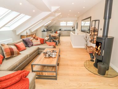 A living room with a large sectional sofa and coffee table facing a wood-burning stove and an open kitchen with a dining table at Sardis Chapel 1 in Malltraeth
