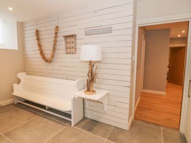 A white wooden bench against a white shiplap wall with a hanging garland and a small shelf holding a lamp next to an open doorway at Sardis Chapel 1 in Malltraeth