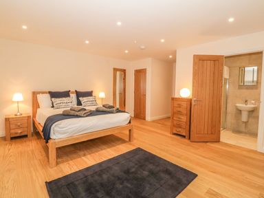 A bedroom with a wooden bed a nightstand with a lamp a dresser a mirror a dark rug and an en suite bathroom at Sardis Chapel 1 in Malltraeth
