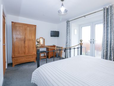 A bedroom with a metal bed, wooden wardrobe, dressing table, TV on the wall, and French doors with curtains at Escape To The Chapel Anglesey in Bryngwran near Rhosneigr