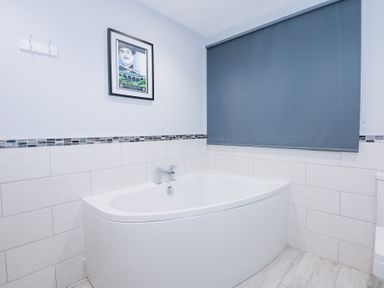 A bathroom with a curved bathtub wall tiles a blue roller blind and a framed picture above the bathtub at Escape To The Chapel Anglesey Bryngwran near Rhosneigr