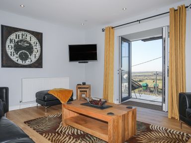 A living room with a wooden coffee table, leather furniture, wall clock, wall-mounted TV, yellow curtains and an open door to a balcony at Escape To The Chapel Anglesey Bryngwran near Rhosneigr