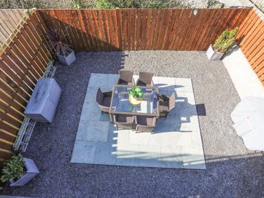 An outdoor patio area with a glass table and six wicker chairs on stone tiles surrounded by gravel and wooden fencing at Escape To The Chapel Anglesey Bryngwran near Rhosneigr