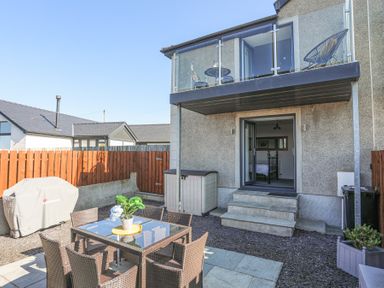 An outdoor patio area with a dining table and chairs a barbecue covered with a cloth a staircase leading to an open door and a balcony with glass railing at Escape To The Chapel Anglesey Bryngwran near Rhosneigr