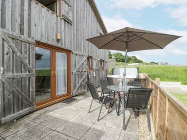 An outdoor patio with a round glass table and four black chairs under a large umbrella and a hot tub beside a wooden house at Top Lodge in Llanvair-Discoed near Penhow