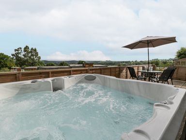 An outdoor area with a bubbling hot tub a table with four chairs and an umbrella with a countryside view at Top Lodge in Llanvair-Discoed near Penhow