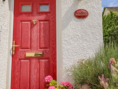 A red door with brass fixtures next to a white wall with a sign that reads The Old Post Office surrounded by flowers and plants at The Old Post Office in Llanelian near Old Colwyn