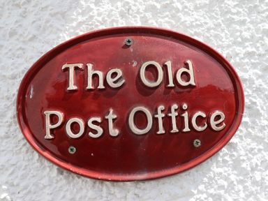 A red oval plaque with the text The Old Post Office mounted on a white textured wall at The Old Post Office in Llanelian near Old Colwyn