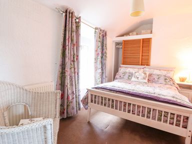 A bedroom with a wooden bed floral bedding curtains wicker chair and bedside table with a lamp at The Old Post Office in Llanelian near Old Colwyn