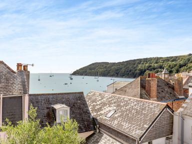 Roofs of houses with chimneys and a view of sailboats on water with a tree-covered hill in the background at Pentreath in Kingsand