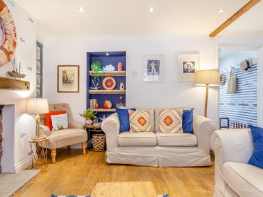 A living room with beige sofas and armchair with cushions bookshelves lamps and framed pictures at Pentreath in Kingsand