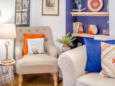 A beige armchair with orange and ship-themed pillows next to a lit lamp and a white sofa with blue and patterned pillows near wooden shelves with books and a vase at Pentreath in Kingsand