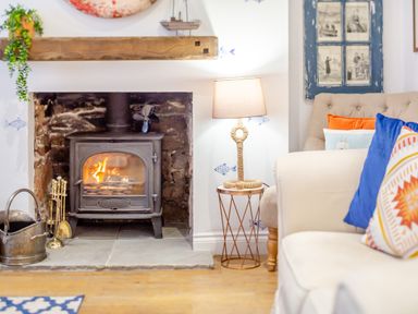 A wood burning stove with fire inside a stone fireplace next to a lamp on a side table and sofas with colorful cushions at Pentreath in Kingsand