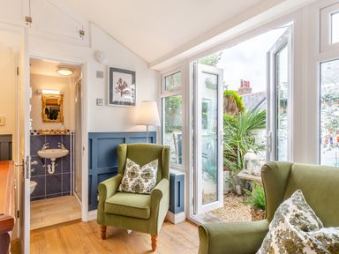 A sitting area with green armchairs and pillows next to open glass doors leading to a garden and a small bathroom with a sink visible at Pentreath in Kingsand