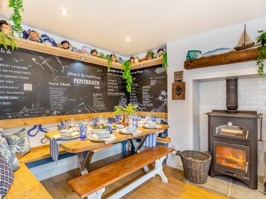 A dining area with a wooden bench table set with blue glasses and white dishes next to a blackboard wall and a wood stove at Pentreath in Kingsand