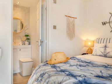 A bedroom with a bed covered by a blue and white blanket a straw hat on the bed a nightstand with a lamp a wall hanging and an open door revealing a bathroom with a toilet and a vanity with a round mirror and plant at Pentreath in Kingsand