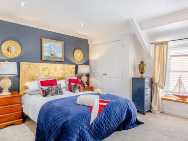 A bedroom with a large bed blue quilt red and floral pillows wooden bedside tables lamps blue dresser and sailboat model at Pentreath in Kingsand