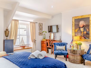 A bedroom with a bed covered by a blue quilt two rolled towels a window with cream curtains a wooden desk with a TV above two blue chairs around a wooden barrel table and a large framed portrait on the wall at Pentreath in Kingsand