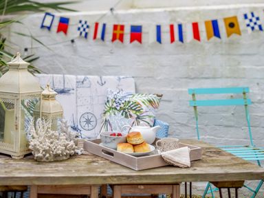 An outdoor wooden table with a tray of scones strawberries a teapot and a cup with lanterns and cushions and a blue chair at Pentreath in Kingsand