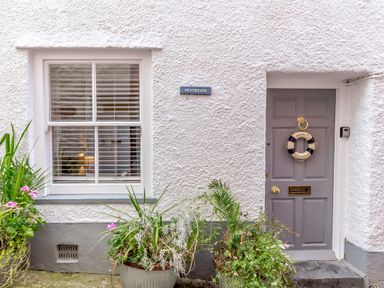 A white textured exterior wall with a window and a grey door decorated with a life ring and plants in pots at Pentreath in Kingsand