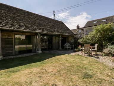 An outdoor garden area with lawn wooden bench and table with chairs next to a stone and wood building at Nympy Cottage in Nympsfield near Nailsworth