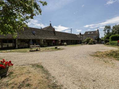A gravel driveway with a wooden picnic table and benches outside a long single-story stone building with a tiled roof at Nympy Cottage in Nympsfield near Nailsworth