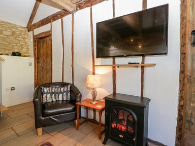 A room with a black armchair a wooden side table with a lamp a black electric fireplace and a wall-mounted tv at Nympy Cottage in Nympsfield near Nailsworth