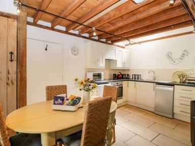 A kitchen with a wooden dining table, woven chairs, white cabinets, and appliances at Nympy Cottage in Nympsfield near Nailsworth