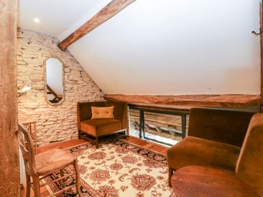 A small attic seating area with two brown chairs a wooden chair a patterned rug a wall mirror and exposed wooden beams at Nympy Cottage in Nympsfield near Nailsworth