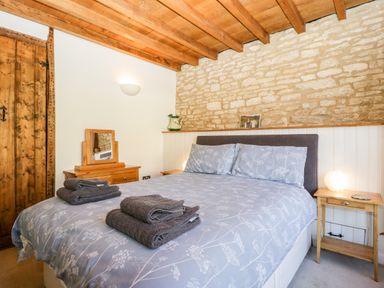 A bedroom with a wooden ceiling a stone wall a bed with grey patterned bedding and folded towels on top at Nympy Cottage in Nympsfield near Nailsworth