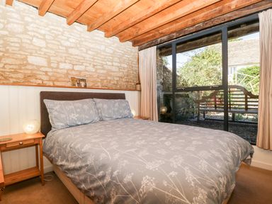 A bedroom with a bed and bedside tables next to a large window showing a bench outside at Nympy Cottage in Nympsfield near Nailsworth