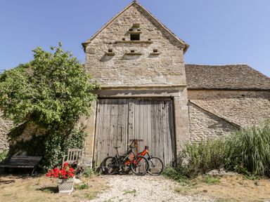 An old stone building with large wooden doors two bicycles parked in front a wooden bench and red flowers in a pot nearby at Nympy Cottage in Nympsfield near Nailsworth