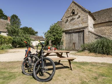Two bicycles parked next to a wooden picnic table with a bottle and glasses outside a stone barn at Nympy Cottage in Nympsfield near Nailsworth