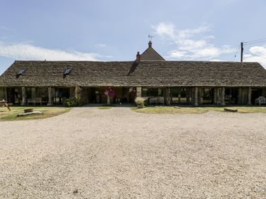 A long single-story stone building with a tiled roof and glass doors with benches and hanging flowers in front at Nympy Cottage in Nympsfield near Nailsworth
