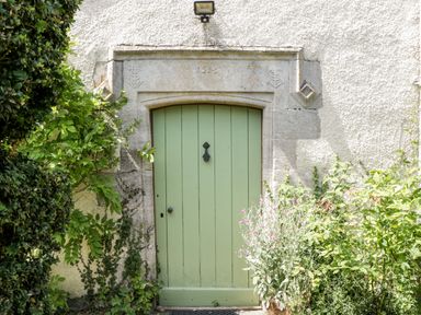 A green wooden door set in a stone frame surrounded by plants and shrubbery at Nympy Cottage in Nympsfield near Nailsworth