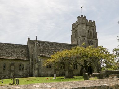 A stone church with a clock tower and a weather vane surrounded by grass and trees at Nympy Cottage in Nympsfield near Nailsworth