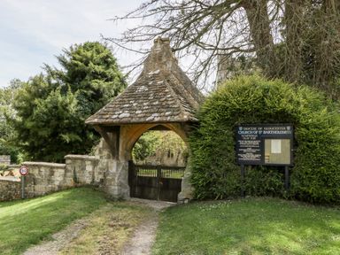 Stone gate entrance with wooden doors and a roof near a signboard and trees at Church of St Bartholomew in Nympsfield near Nailsworth
