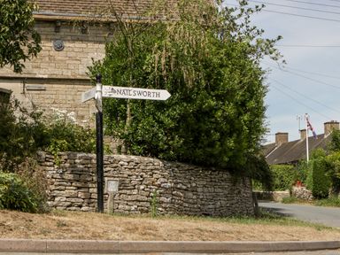 A road sign pointing to Nailsworth and Stroud near a stone wall and bush in a rural village