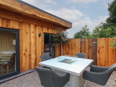 An outdoor patio with a white table with a glass fire feature and four gray chairs next to a wooden building and fence at Daffodil in Llangadfan