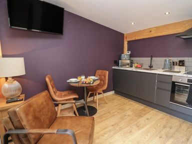 A kitchen area with a small round table and two chairs a brown armchair a wall-mounted TV and kitchen cabinets with appliances at Daffodil in Llangadfan