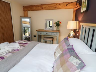 A bedroom with a bed with plaid pillows and a blanket a dressing table with a mirror and flowers a floor mirror and a lamp at Daffodil in Llangadfan