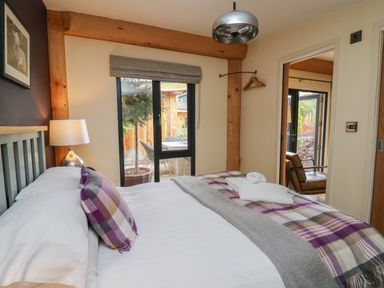 A bedroom with a bed with plaid pillows and a throw blanket a lamp on a bedside table and a view into a room with a brown armchair at Daffodil in Llangadfan