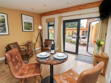 A dining area with a small round wooden table set for two wine glasses and chairs near glass doors opening to a garden at Rhosyn in Llangadfan
