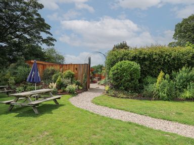 A garden with a wooden picnic table with an umbrella a gravel path and green bushes at Rhosyn in Llangadfan