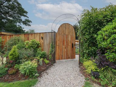 A wooden garden gate with a metal arch on a gravel path surrounded by plants and shrubs at Rhosyn in Llangadfan