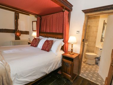 A bedroom with a four-poster bed with red canopy and pillows and a bedside table with a lamp next to a bathroom with patterned floor tiles at Pentre Hall in Bronygarth near Chirk